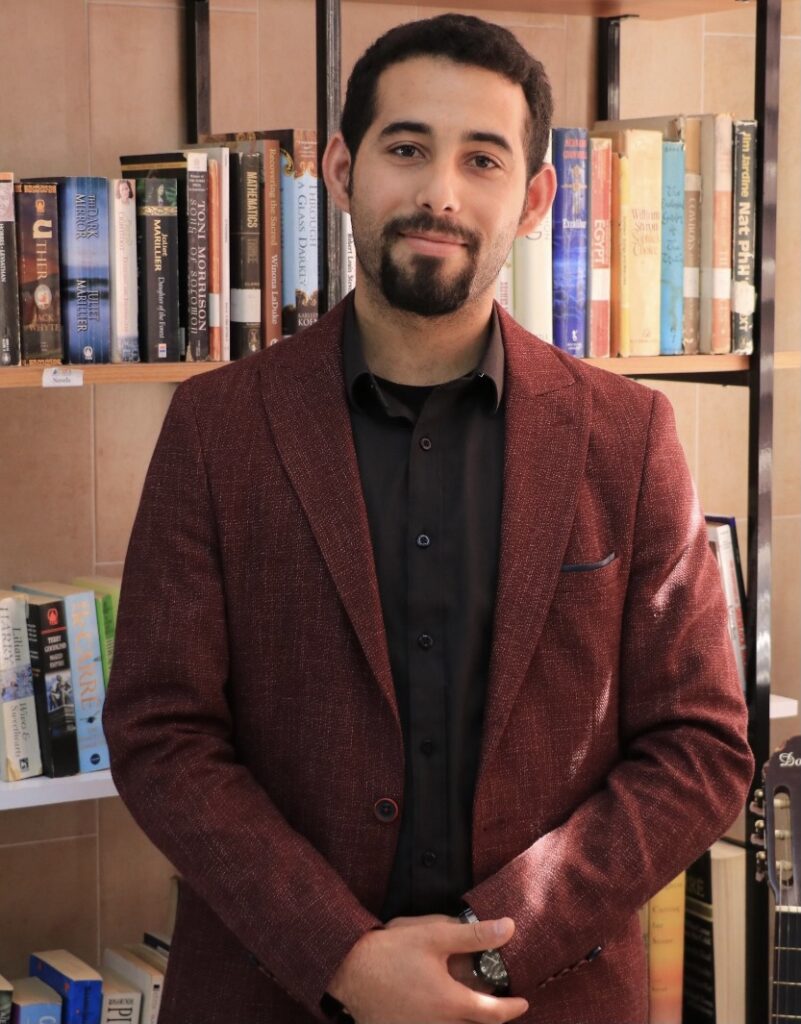 Mosab Abu Toha standing in front of a bookshelf with hands crossed in front