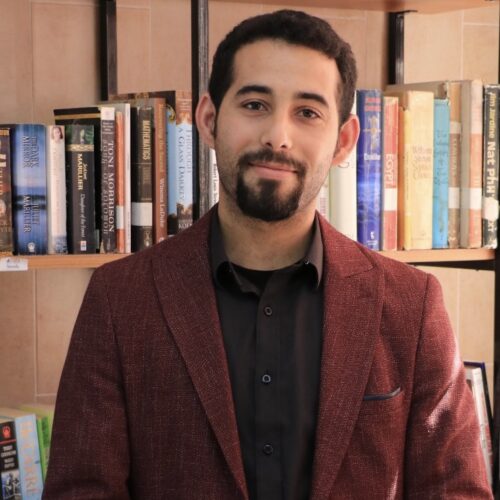 Mosab Abu Toha standing in front of a bookshelf with hands crossed in front